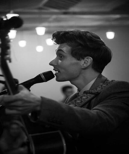 Black and white image of a person stood next to a microphone singing ad playing a guitar