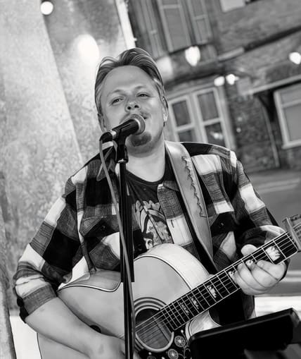 Black and white image of a person sat next to a microphone singing ad playing a guitar