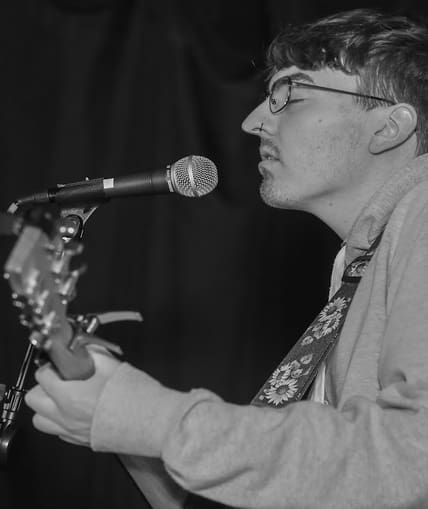 Black and white image of a person stood next to a microphone singing ad playing a guitar