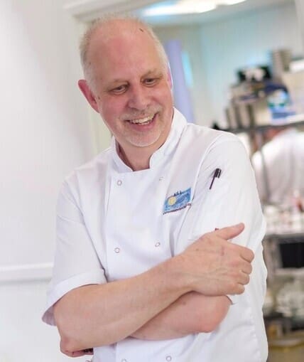 A person stood in chef whites with their arms crossed and a kitchen in the background.